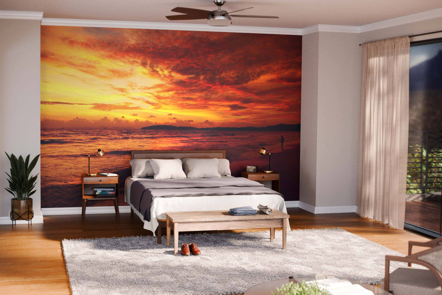 A modern bedroom features a large bed, wooden furniture, and a gray rug. Behind the bed, the Sea Water Wall Mural adds warmth with its orange and yellow ocean sunset design. Sunlight filters in through sheer beige curtains over tall windows.