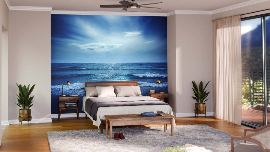 A modern bedroom showcases the Sealion Seascape Wall Mural behind the bed. White bedding, two nightstands, plants, a wooden bench, and a soft rug complete the look. Sunlight streams in through floor-to-ceiling glass doors with beige curtains.