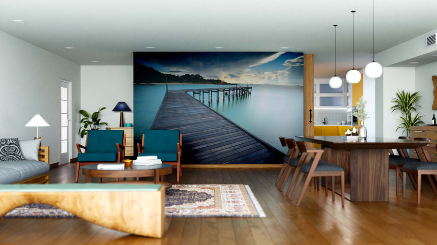Modern open-concept living and dining room with wooden floors, blue armchairs, Seascape Wall Mural of a serene pier over water, pendant lights, rugs, green plants, and a kitchen in the background.