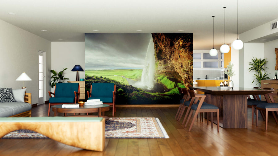 A modern open living and dining room features wood flooring, stylish blue and wooden furniture, a rug, plants, pendant lights, and the Seljalandfoss Waterfall Summer Time Wall Mural as a natural focal point.