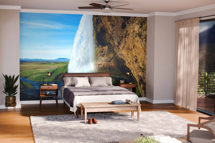 A cozy bedroom with wood furniture and neutral bedding features the Seljalandsfoss Waterfall Iceland Wall Mural, creating an illusion of an open window to nature. Large windows bring light in; a rug and bench are at the foot of the bed.