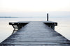 The Serene Wooden Dock Wall Mural captures a weathered pier extending into calm waters under an overcast sky, with a solitary post at its end. Faint outlines of distant land on the horizon and the sky's reflection enhance the mural's tranquil atmosphere.