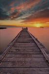 Serenity at Dusk Wall Mural features a weathered pier stretching into calm water under a vibrant sunset. Orange, yellow, and pink hues reflect on the sea—ideal as a custom printed or peel and stick mural to bring tranquility to any space.