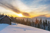 A rustic wooden cabin in a snowy forest is illuminated by sunrise, as dramatic clouds streak across the sky. Distant mountains complete this serene winter view, reminiscent of the "Severe Winter on a Mountain Range Wall Mural.