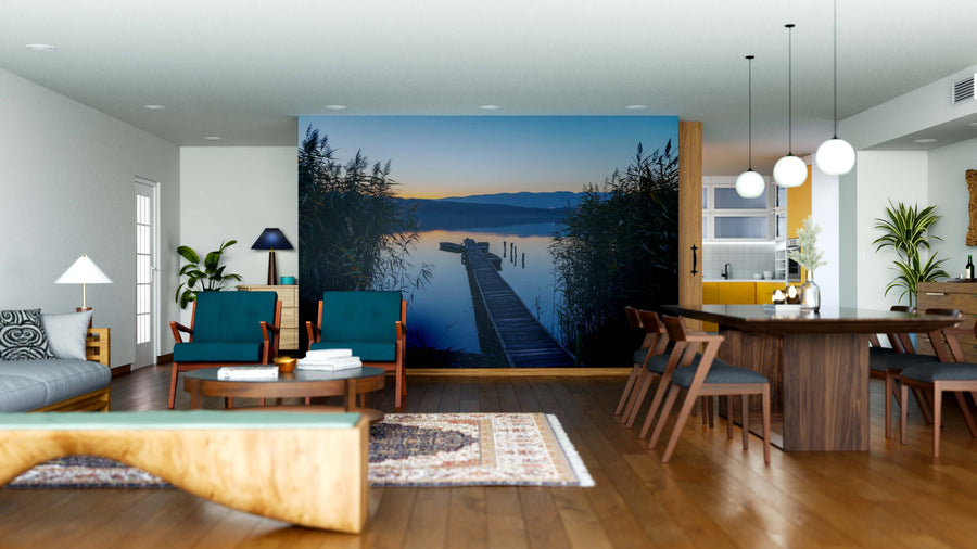 A modern open-plan living and dining area features wooden furniture, a rug, and the Silent Lake Wall Mural depicting a sunset lake with dock, reeds, and mountains. Pendant lights hang above the dark dining table with six chairs for a tranquil vibe.