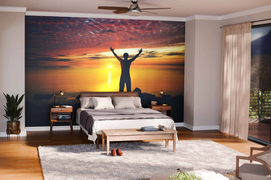 A cozy bedroom showcases the Silhouette of a Person Wall Mural—a striking image of a figure with raised arms against a sunset behind the bed—enhanced by warm lighting, wooden furniture, plants, and large windows with sheer curtains.