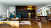 A modern living-dining room features wood furniture, blue chairs, and the Silhouetted Pier Glow Wall Mural depicting a sunset over water. Pendant lights, rugs, plants, and a blend of contemporary and rustic decor make the space inviting.