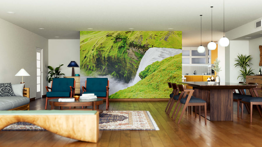 A modern open-plan living and dining area features wood floors, stylish furniture, and the Skogafoss Waterfall Wall Mural, bringing lush natural scenery indoors. Pendant lights hang above the dining table as natural light brightens the space.