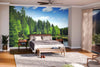 A modern bedroom with a wooden bed, nightstands, plants, and a bench. The main wall features the Arber Lake Germany Wall Mural, depicting a lush forest and lake. Sunlight enters through sheer curtains and a glass door.