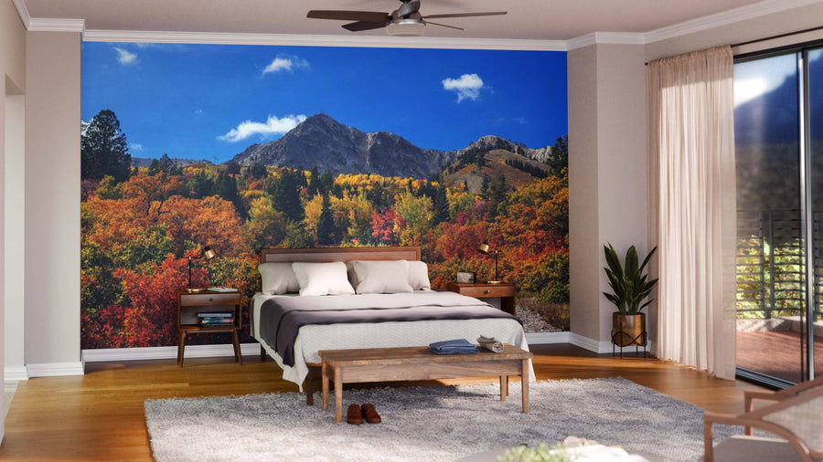 A bright bedroom features the Snow Basin Utah Mountains Wall Mural of autumn trees and peaks behind the bed, along with a modern bed, neutral bedding, wooden nightstands, a bench, gray rug, potted plant, and sunlit sliding glass doors.