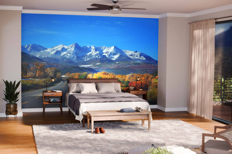 A bright bedroom features the Snow on Dallas Divide Mountains Wall Mural behind a bed with beige and brown bedding. Wooden nightstands, a bench, a light rug, potted plant, and sunlight from glass doors complete the cozy scene.