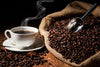 The "Steaming Hot Coffee Wall Mural" by Photo features a steaming cup of black coffee on wood, surrounded by a burlap sack spilling roasted beans and a metal scoop, captured in warm light to enhance the rustic atmosphere.