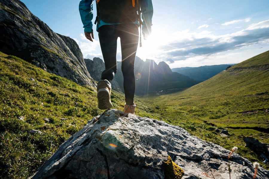 A modern living-dining room with wooden floors, green and teal furniture, and pendant lights features the striking Steps to Summit Wall Mural, showing a close-up of a hiker’s legs atop a rock in a sunlit mountain landscape.