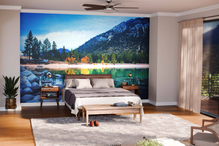 A bright bedroom with wood floors, gray and white bedding, nightstands, lamps, and a bench. The back wall displays the Still Water Lake Tahoe Wall Mural, creating a serene scene as sunlight streams through sheer curtains.