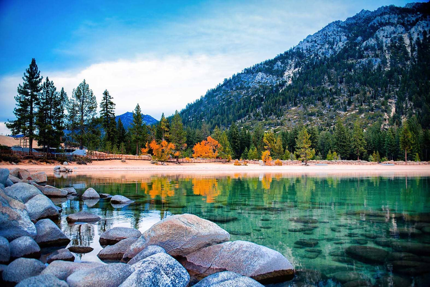 Transform your space with the Still Water Lake Tahoe Wall Mural, featuring a serene landscape of a clear lake, large rocks, and vibrant autumn trees reflecting on the water. In the background, pine trees and a mountain stand tall under a partly cloudy blue sky.