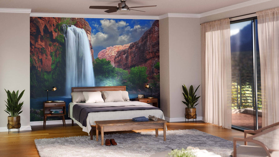 A modern bedroom showcases a Stunning Waterfall Wall Mural behind a wooden bed, with matching nightstands, lamps, two potted plants, a bench, light curtains, ceiling fan, and glass doors that lead to a balcony with lush greenery.