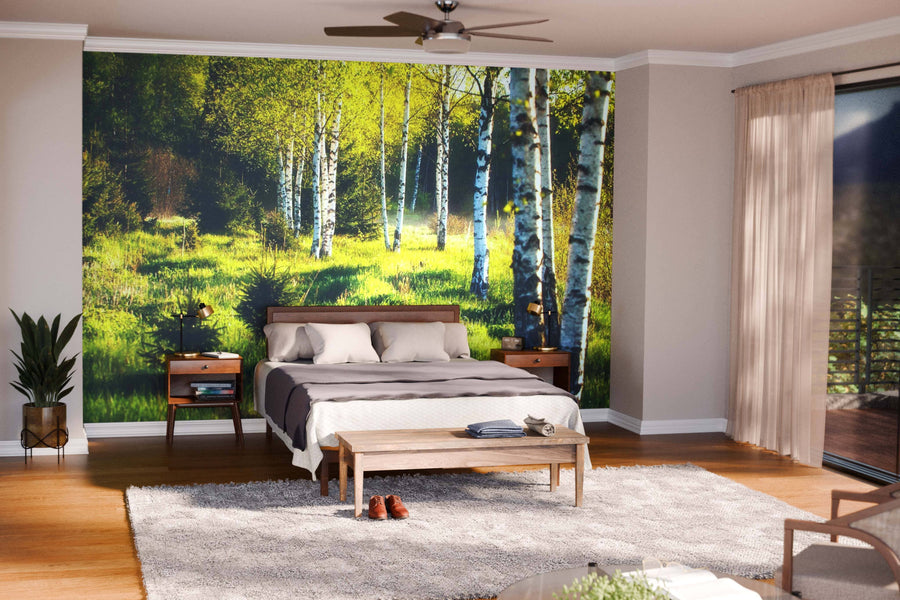 A cozy bedroom with a large bed, wooden furniture, and a light rug. The back wall displays the Summer Forest In Sunlight Wall Mural, bringing a nature-inspired vibe as sunlight streams through sheer curtains into the inviting room.