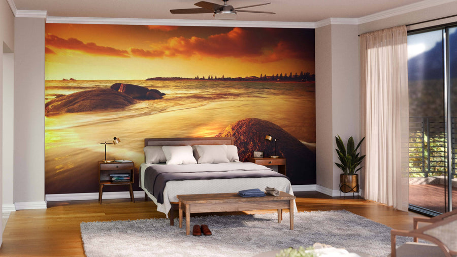 A modern living-dining room features wooden floors, mid-century furniture, and a Sun Tinted Beach Wall Mural of an orange sunset over ocean rocks. Pendant lights hang above a dark dining table, while cozy rugs add warmth to the inviting space.