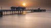The Sunbound Wall Mural depicts a serene scene of a long pier stretching into calm waters at sunrise, where the sun casts an orange glow, silhouetting buildings and their gentle reflections—perfect inspiration for a peel-and-stick wall mural.