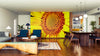 A modern living-dining room features wood floors, blue chairs, a gray sofa, and a large Sunflower Core Wall Mural. A long dining table with wooden chairs sits under globe lights, with the kitchen visible in the background.