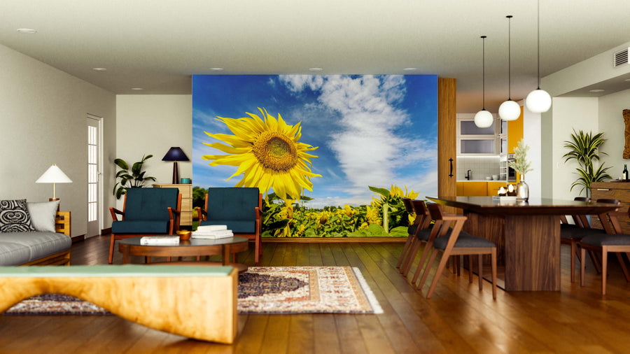 A bright living-dining room with wood floors and mid-century furniture features the Sunflower Under Sky Wall Mural as a striking accent wall. Plants, rugs, and pendant lights enhance the cozy, inviting atmosphere.