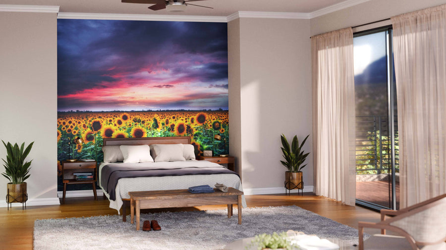 A bright, modern bedroom features the Sunflower on Sunset Wall Mural behind the bed, flanked by two nightstands, green plants, floor-to-ceiling windows with sheer curtains, a wooden bench at the foot of the bed, and a balcony outside.