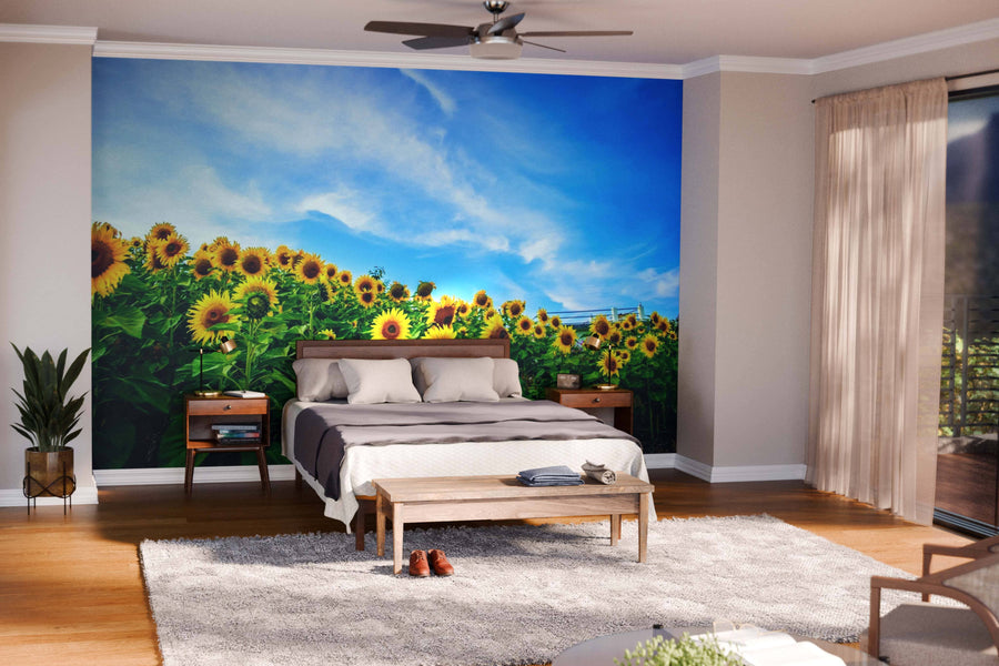 A modern bedroom showcases the "Sunflower on the Hill Wall Mural" behind a wooden bed with beige bedding. Bedside tables hold lamps and plants, while a soft rug, bench, and shoes rest on the floor. Sunlight pours in from glass doors.