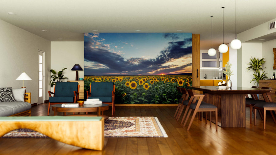 A modern living-dining room features wooden floors, contemporary furniture, and the "Sunflowers at Dusk Wall Mural," displaying a sunflower field at sunset. Warm lighting, rugs, and potted plants enhance the cozy atmosphere.
