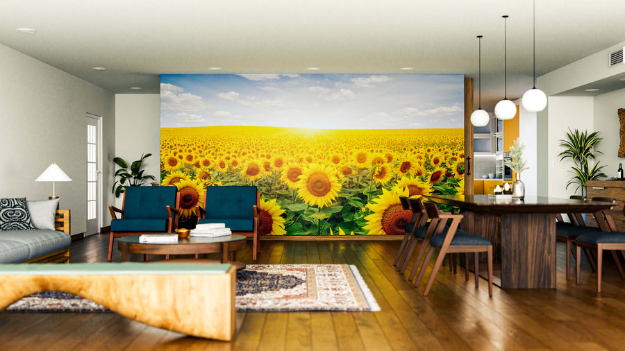 A modern living-dining room showcases the Sunlit Flower Field Wall Mural, a large, vibrant peel and stick decor featuring a sunflower field at sunset. The space also includes wood flooring, blue armchairs, a wooden dining table, and pendant lights.