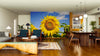 Modern living and dining room with wooden furniture, rugs, and plants. The Sunny Day Flower Wall Mural featuring a vibrant yellow sunflower and blue sky adds bright color to the neutral space. Pendant lights hang above the dining table.