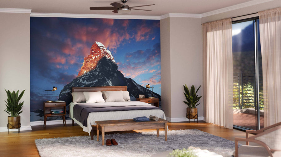 A modern bedroom features the Matterhorn Mountain Swiss Alps Wall Mural behind the bed. Beige walls, wood floors, minimalistic furniture, and two plants complete the serene and stylish space.