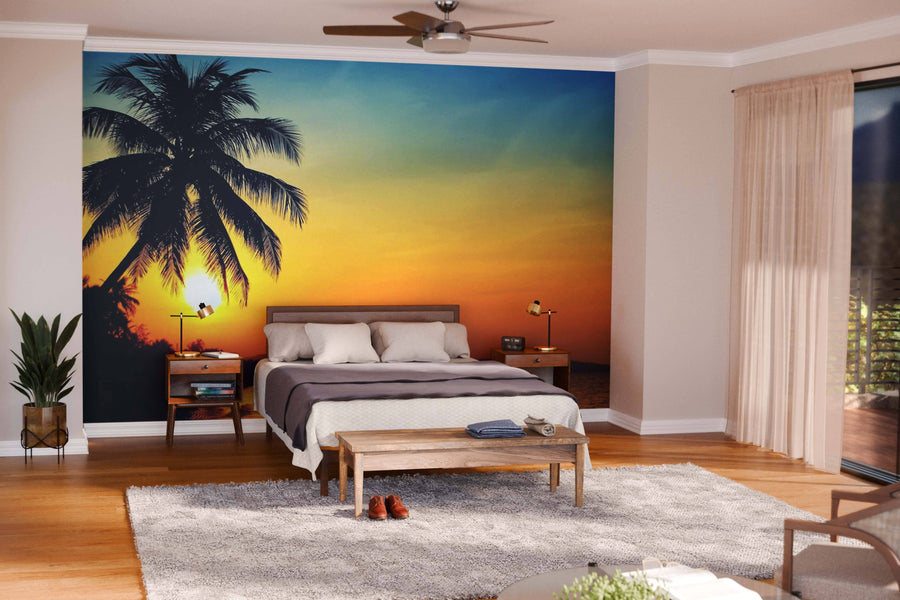 Modern living-dining room with wooden floors, teal chairs, a rug, and a large Sunrise in the Tropics Wall Mural featuring a palm tree over the ocean; pendant lights hang above the dark dining table with the kitchen visible behind.