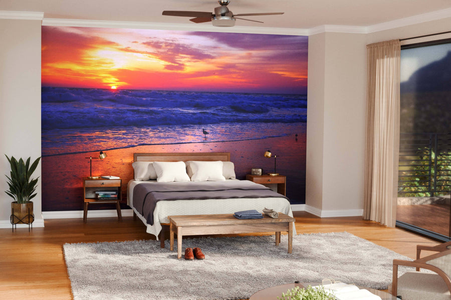 A modern living-dining room features wooden floors, stylish furniture, pendant lights, and a wall-sized Sunrise on the Surf Wall Mural, showcasing a vibrant ocean sunrise that adds warmth and color to the open-concept space.