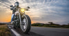 Wearing a black helmet and jacket, the motorcyclist rides a classic bike on an open road at sunset in this "Sunset Biker Wall Mural," where the headlight beams through an orange and blue sky with wispy clouds framing the scene, epitomizing speed and freedom against grassy hills.