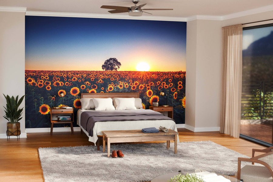 A cozy bedroom with wooden furniture features the Sunset Blooming Over Sunflower Field Wall Mural behind the bed. Sunlight from large windows highlights the inviting wooden floor and warm decor.