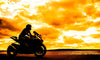 A silhouetted motorcyclist rides under a vibrant orange and yellow sunset sky, evoking the essence of the "Sunset Chase Wall Mural." Dramatic clouds and a body of water enhance the striking contrast between the dark silhouette and the bright, mural-like horizon.