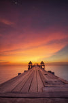 The Sunset Over Pier Wall Mural by features a wooden pier leading to a gazebo-like structure set against a vibrant sunset sky, with orange, pink, and purple hues dotted with stars, capturing nature's serene beauty reflected in the calm water below.