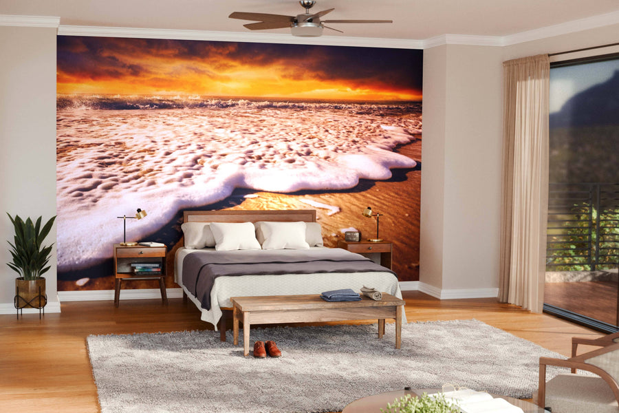 A modern bedroom showcases the Sunset Shore Wall Mural behind the bed, complemented by neatly made bedding, wooden furniture, houseplants, and glass doors leading to a balcony for a serene atmosphere.