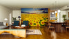 A modern living and dining room with wooden floors and stylish furniture features the Sunset Symphony Wall Mural, showcasing a sunflower field at sunset. Warm lighting and a cozy atmosphere complete this stunning space.