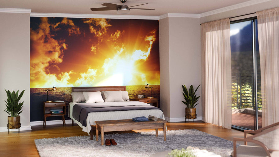 A modern bedroom showcases a bed with white and gray bedding, two wooden nightstands, and the Sunset and Sea Wall Mural creating a vibrant ocean sunset focal point. Large windows with sheer curtains brighten the space, while two potted plants flank the bed.