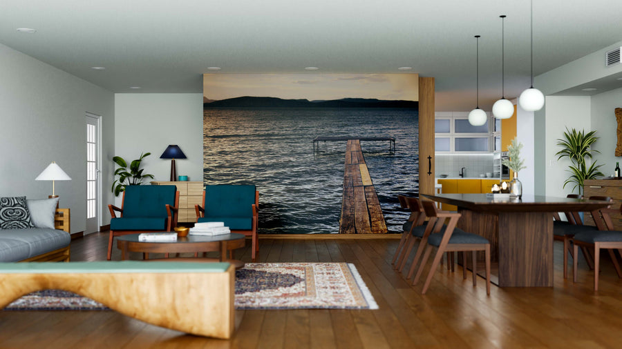 A modern open living-dining area with hardwood floors, blue chairs, a wooden dining table, and kitchen is highlighted by the Sunset at Lake Wall Mural—a serene mural of a calm pier at sunset that serves as a stunning focal point.