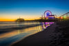 Experience the "Sunset at the Pier Wall Mural", showcasing a vibrant beach scene with a glowing Ferris wheel. The sky shifts from deep blue to orange, reflecting on water and wet sand, akin to removable wallpaper filled with colorful lights and a serene shoreline.