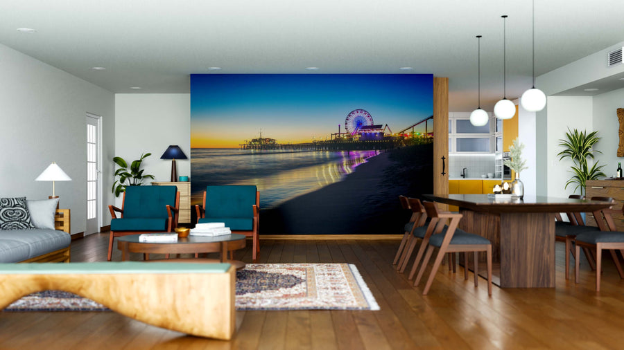 A modern living-dining room with wooden floors and stylish furniture features the "Sunset at the Pier Wall Mural," depicting a lit-up pier, Ferris wheel, and calm ocean at dusk for a vibrant coastal touch in the open, airy space.