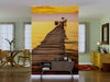 A modern living room with light green walls features the "Sunset on the Pier Wall Mural," showcasing a wooden pier at sunset over tranquil water. Mid-century furniture and plants add warmth and style to the inviting space.