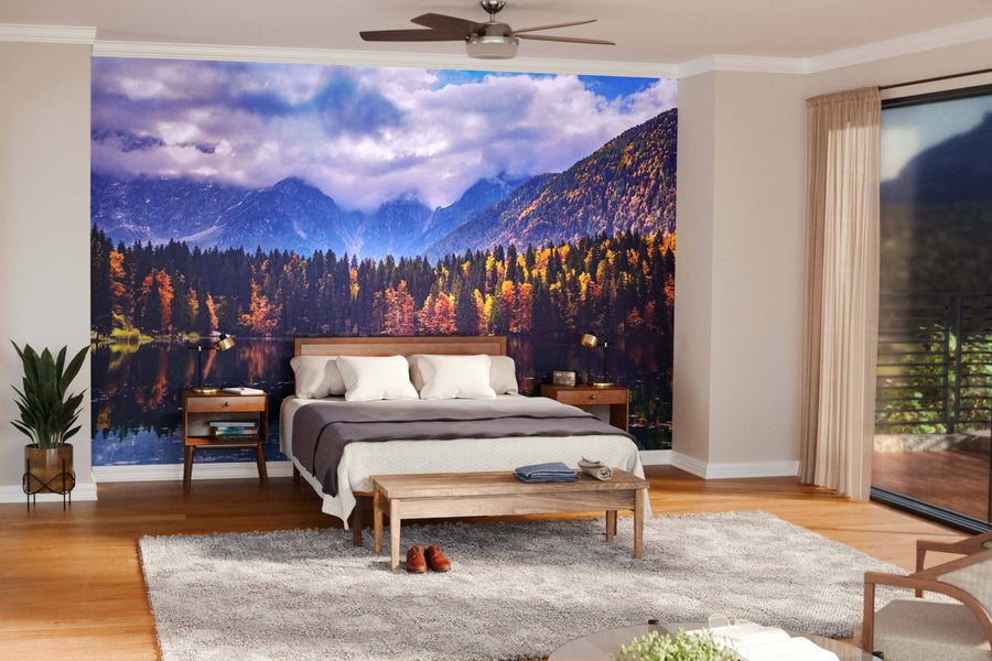 A cozy bedroom showcases a bed with white and beige bedding, wooden nightstands, plants, and the "Swans on Lake Fusine Wall Mural" depicting a scenic lake. Large windows brighten the room, and shoes sit on a beige rug.
