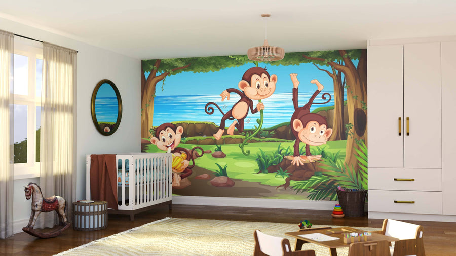 A bright nursery features the Swinging Monkeys Wall Mural with three cartoon monkeys in a jungle by water, along with a crib, rocking horse, basket, toys, white furniture, and sunlight through large windows with sheer curtains.