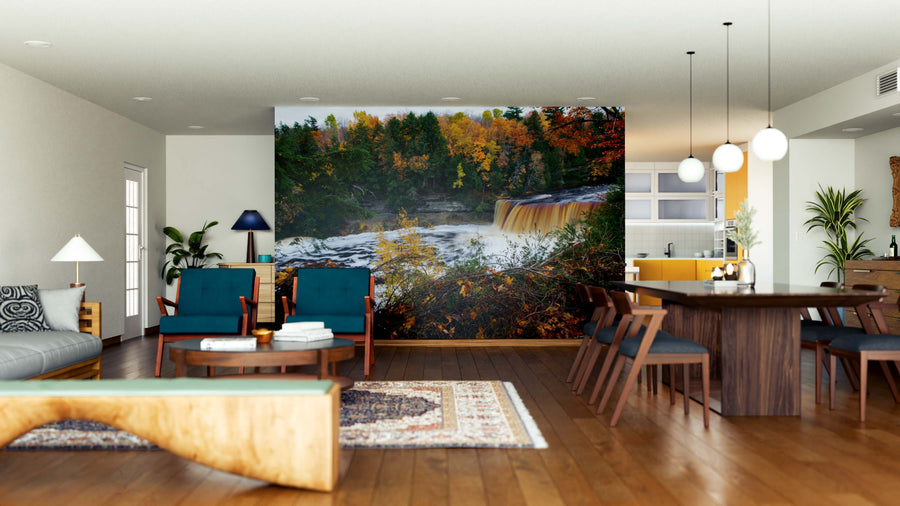 A modern open-plan living and dining area features wooden floors, teal chairs, and the Tahquamenon Falls Wall Mural showcasing a forest waterfall amid autumn foliage. Plants, pendant lights, and a patterned rug complete the decor.
