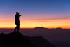 A silhouette of a person in a beanie and jacket stands on a mountain peak, pointing towards distant ranges. The scene resembles the Thumbs Up Wall Mural  with blues and oranges suggesting sunrise or sunset, capturing nature's grandeur as clouds hover below.