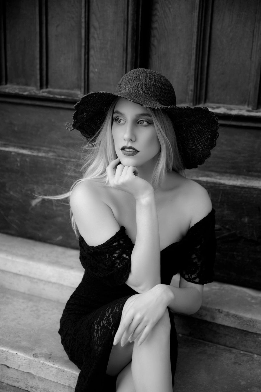 A woman in a black off-the-shoulder dress and wide-brimmed hat sits on stone steps, thoughtfully gazing aside. Behind her, a wooden door stands out like the perfect backdrop for the Timeless Elegance Wall Mural.