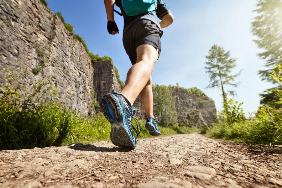 A modern living-dining room with wooden floors, teal chairs, and pendant lights features the Trail Run Wall Mural—an easy-to-update, peel-and-stick wallpaper showing a runner’s legs on a scenic trail amid greenery and stone ruins.